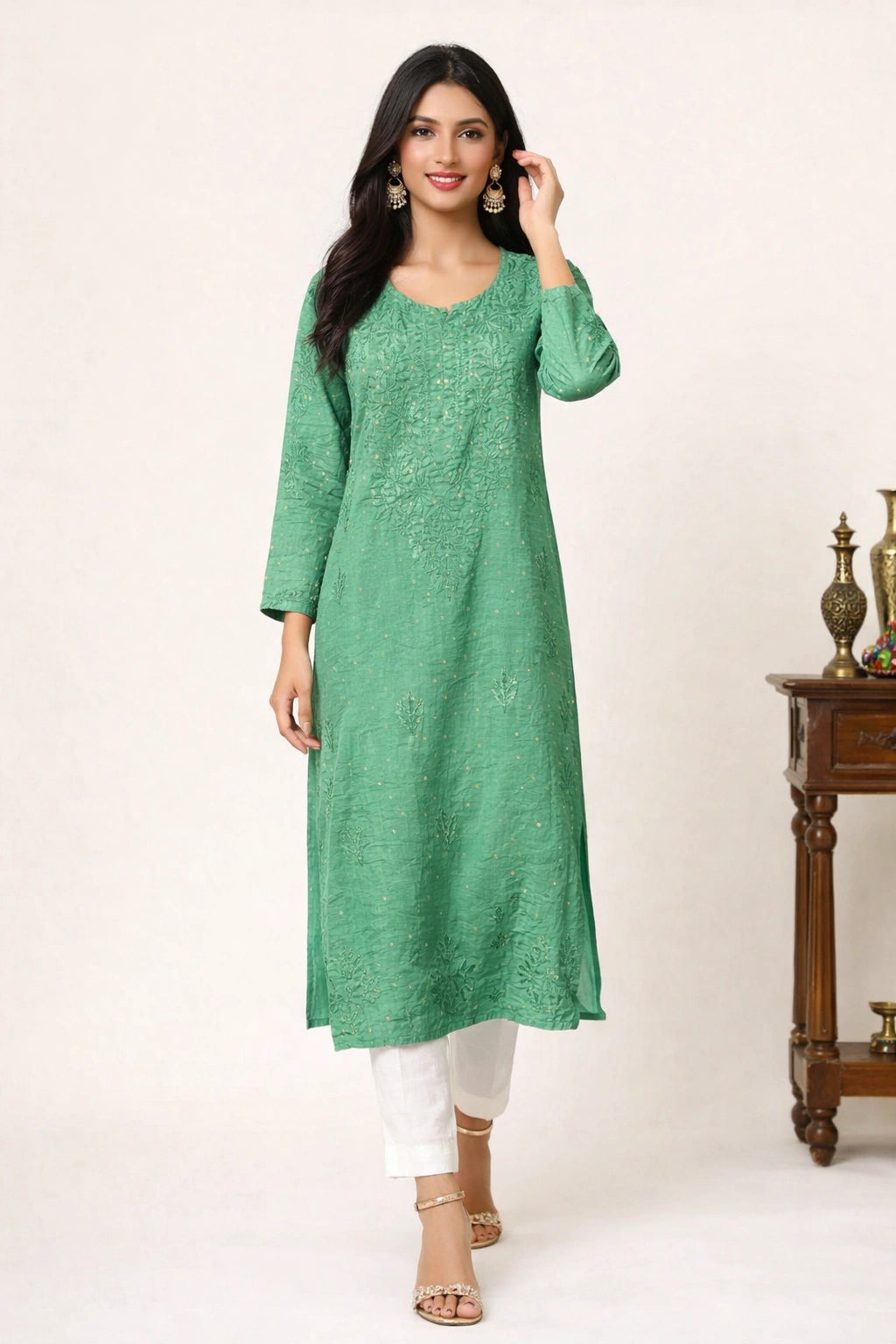 A woman modeling a green embroidered kurta with white pants and gold sandals, standing in a studio setting with a wooden side table visible on the right.