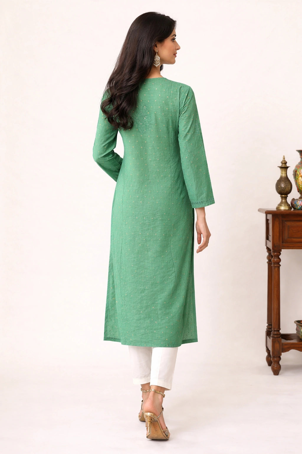 Lucknowi chikankari A woman wearing a sage green long-sleeve kurta with white pants and gold sandals, shown from the back in a studio setting with a wooden side table in the background.