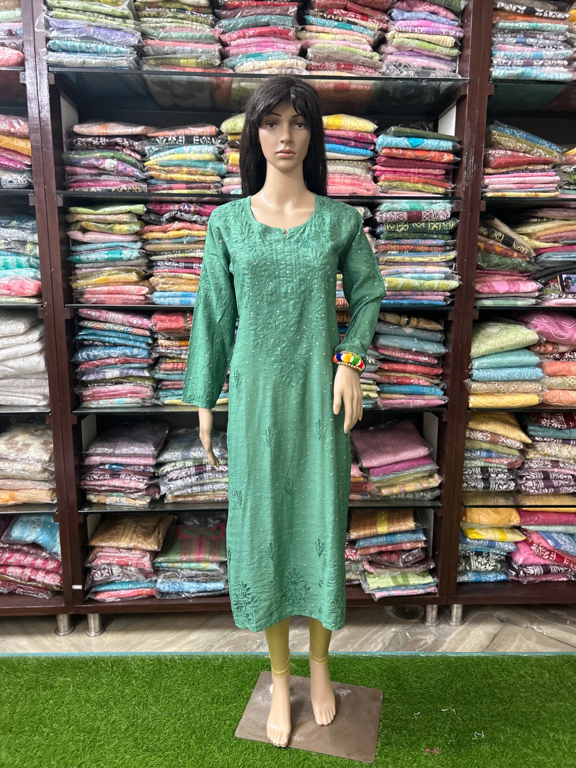 A mannequin wearing a long-sleeved green embroidered kurta stands in front of shelves filled with folded colorful fabrics.