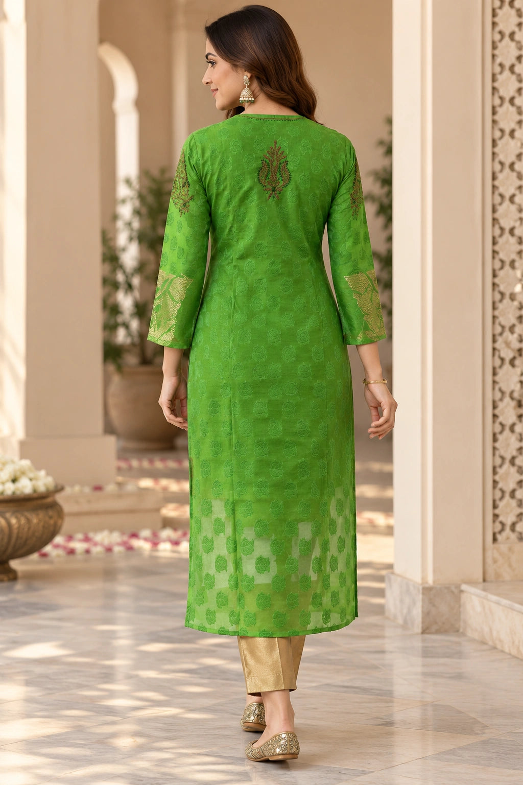 A woman wearing a bright green embroidered kurta with gold-patterned pants, shown from the back in an elegant indoor corridor.