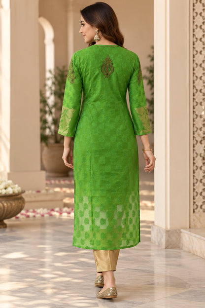 A woman wearing a bright green embroidered kurta with gold-patterned pants, shown from the back in an elegant indoor corridor.