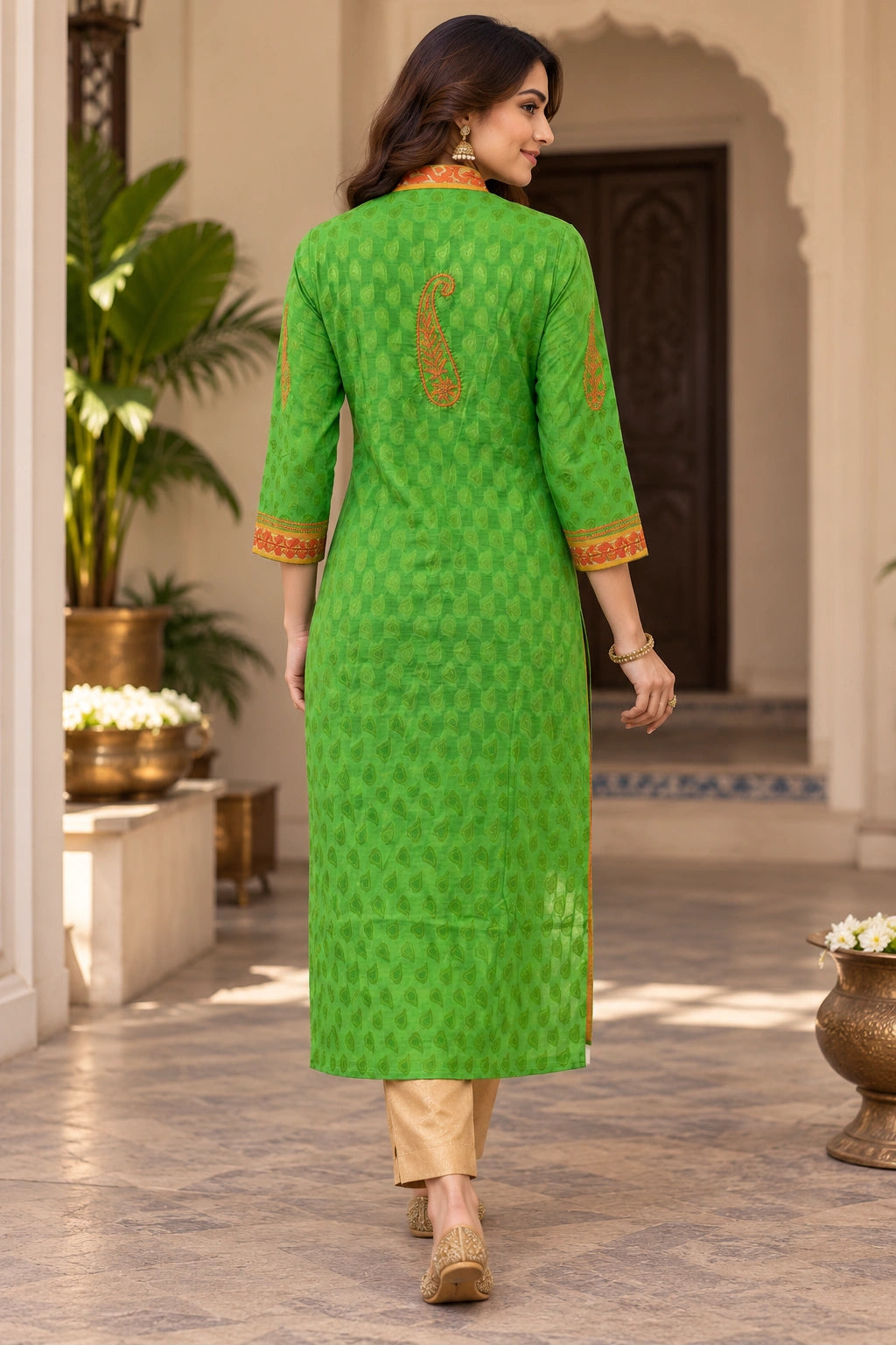 Lucknowi chikankari A woman wearing a bright green kurta with gold and orange patterned details, paired with beige trousers, walking in a courtyard.