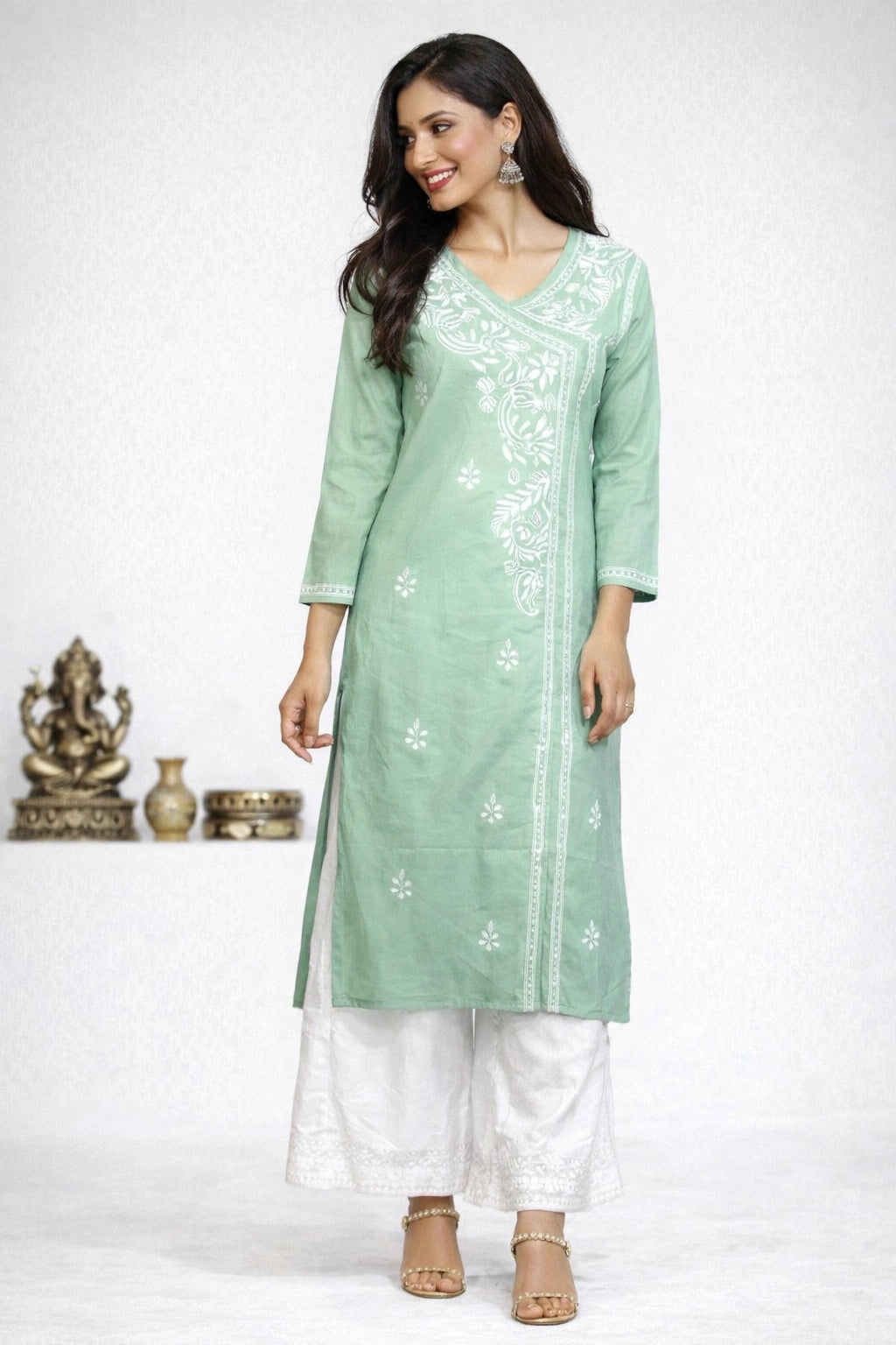 A woman modeling a mint green kurta with white floral embroidery, paired with white palazzo pants and beige sandals, set against a white studio background with decorative brass items on the left.