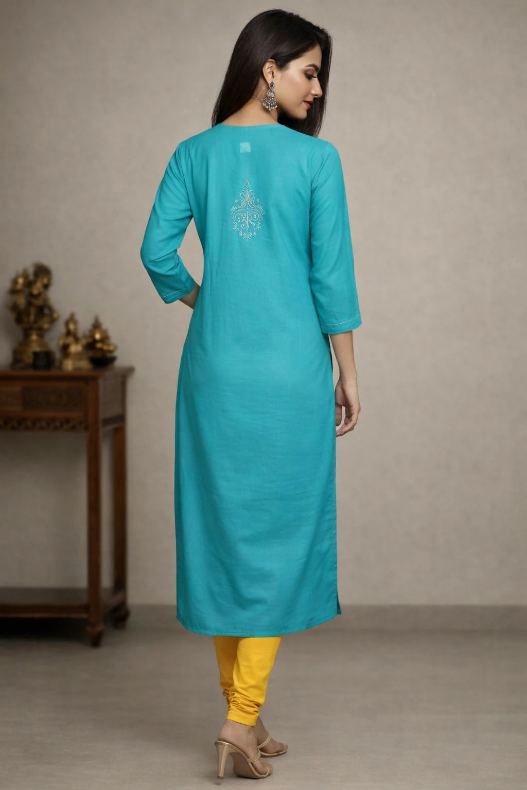 A woman wearing a turquoise blue three-quarter sleeve kurta with silver embroidery on the back, paired with yellow pants and beige heels, shown from a rear three-quarter view in an indoor setting.