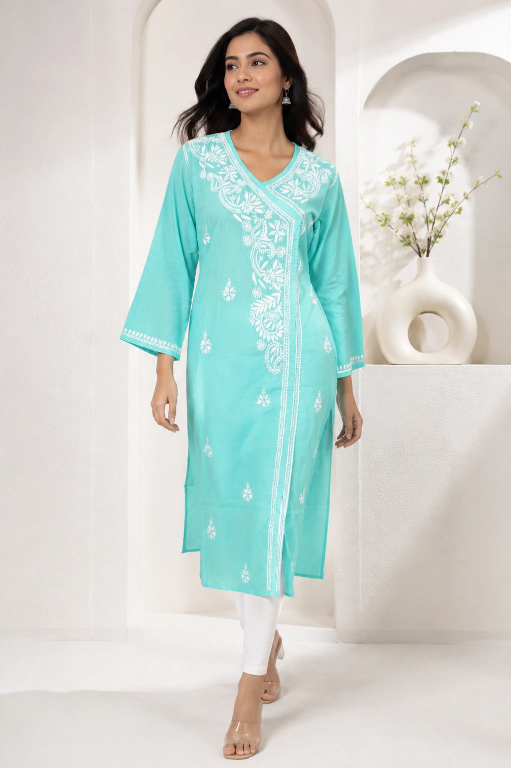 A woman modeling a turquoise mint green kurta with white embroidery, paired with white pants and clear high-heeled sandals, set against a minimalist white architectural background.