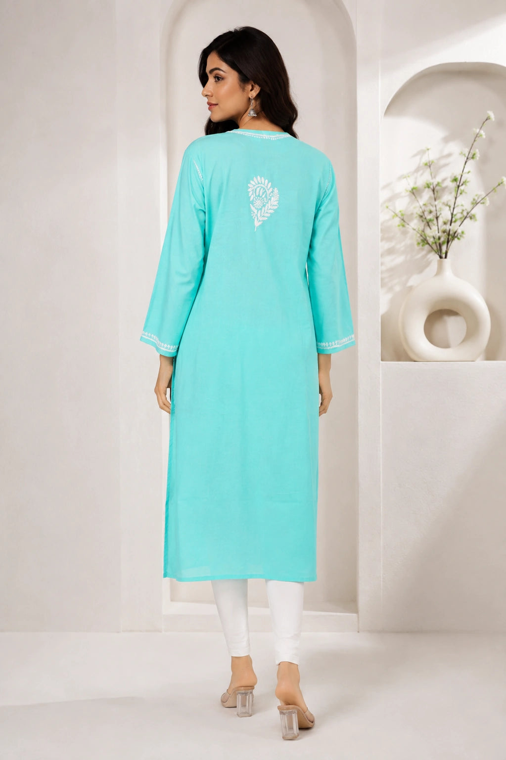 A woman modeling a light turquoise long kurta from the back, featuring a white embroidered heart design on the upper back and white piping on the sleeve cuffs, paired with white leggings and clear high-heeled sandals in a minimalist white interior setting.