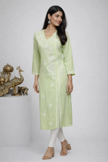 A woman modeling a light green kurta with white floral embroidery, paired with white pants and beige sandals, standing against a light gray studio background with a decorative gold peacock statue on the left.