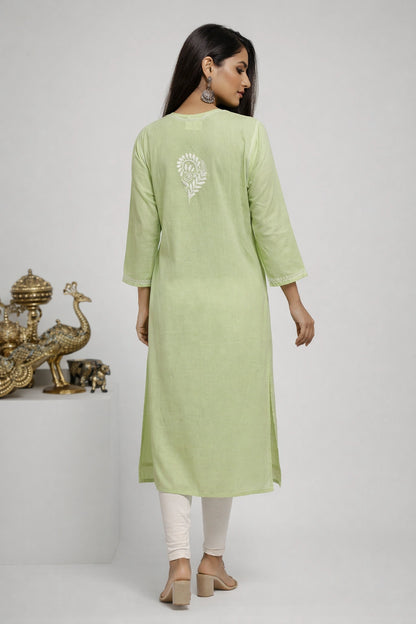 A woman is shown from the back wearing a light sage green kurta with a white embroidered motif on the upper back, paired with white leggings and beige block-heel sandals, set against a neutral studio background with decorative gold peacock figurines on the left.
