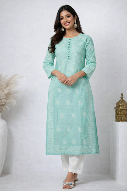  Lucknowi chikankari A woman modeling a mint green kurta with white printed motifs and white pants, standing in a studio setting with decorative pampas grass and a brass 