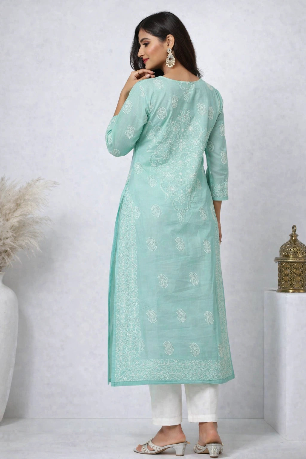 Lucknowi chikankari A woman modeling a mint green kurta with white printed patterns, paired with white pants and silver sandals, shown from the back against a textured light gray wall.