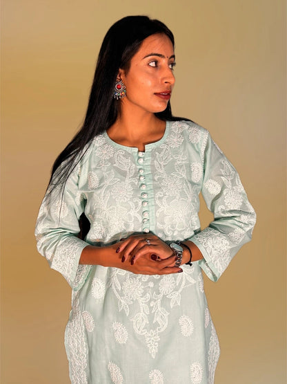 Lucknowi chikankari A woman modeling a light sage green kurta with white floral embroidery and a buttoned neckline, set against a solid beige background.