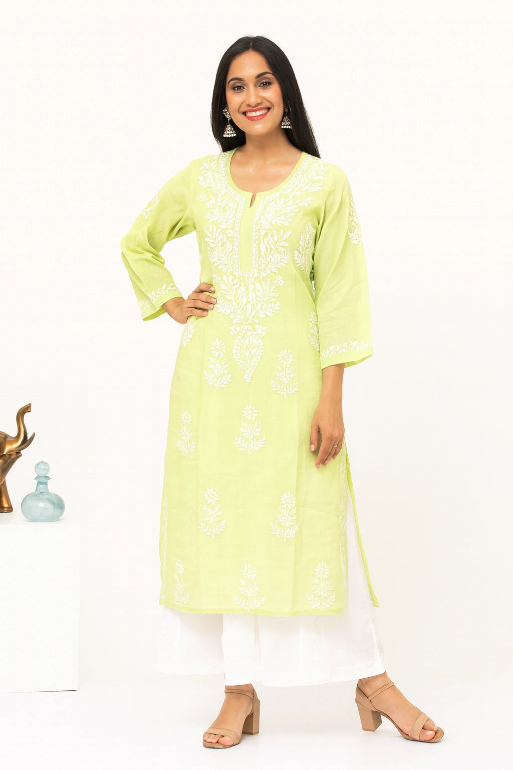 A woman modeling a light yellow-green kurta with white floral embroidery, paired with white pants and beige heeled sandals, set against a plain white studio background.