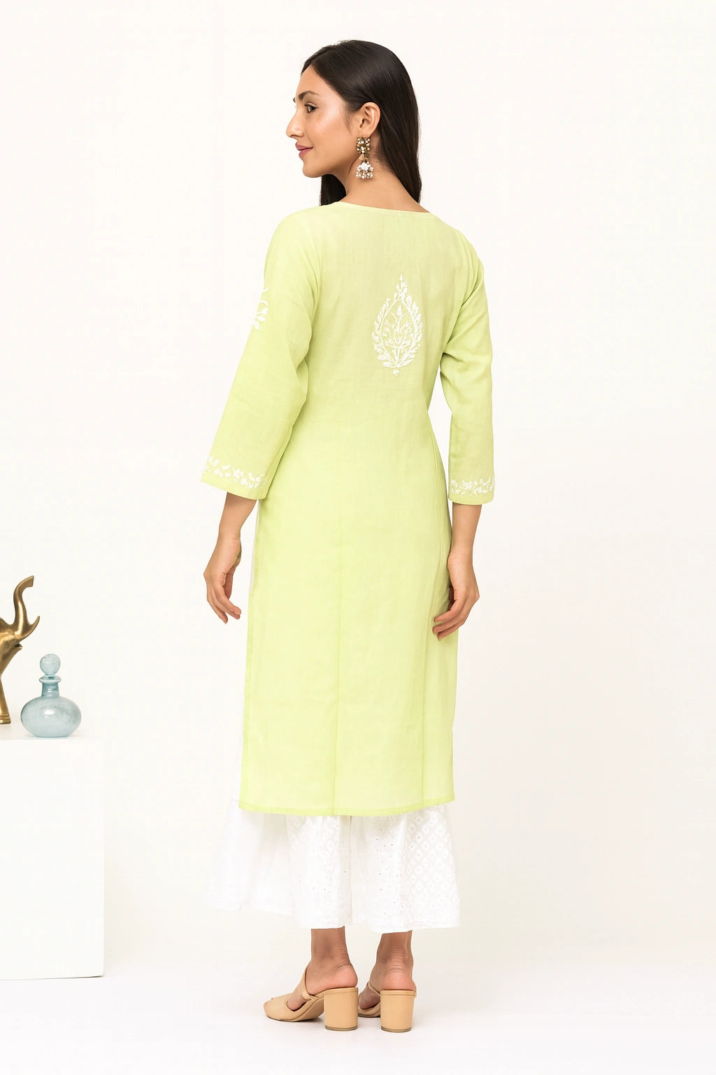 Lucknow chikankari A woman modeling a light yellow-green kurta from the back, featuring white embroidery on the back and cuffs, paired with a white flared skirt and beige block-heel sandals against a white studio background.