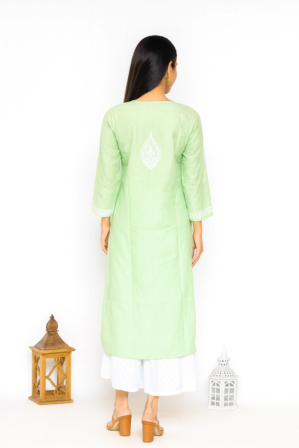 A woman viewed from the back wearing a light green long kurta with white embroidery on the upper back, paired with white pants and tan heeled sandals, standing against a white studio background with two lantern props.