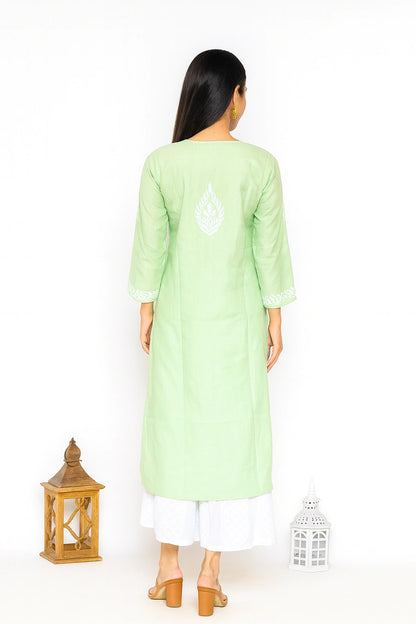 A woman viewed from the back wearing a light green long kurta with white embroidery on the upper back, paired with white pants and tan heeled sandals, standing against a white studio background with two lantern props.