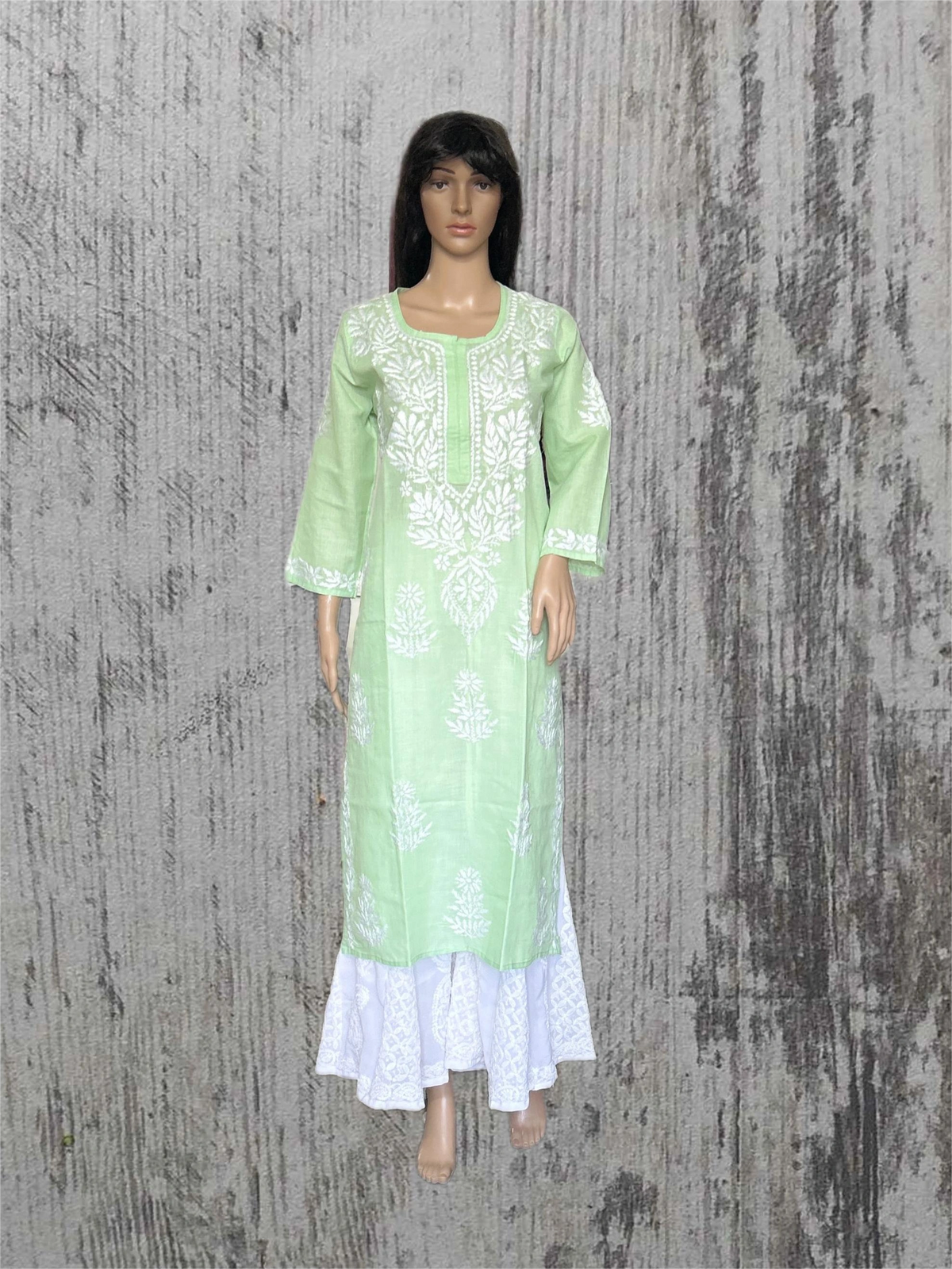 A light green long kurta dress with white floral embroidery and a white lace hem, displayed on a female mannequin against a textured gray background.
