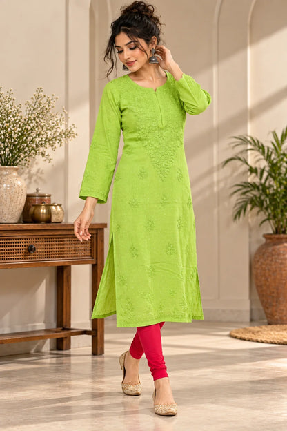 A woman models a bright lime green embroidered kurta with three-quarter sleeves, paired with bright pink leggings and beige flat shoes, standing in a styled indoor room with plants and a wooden console table.