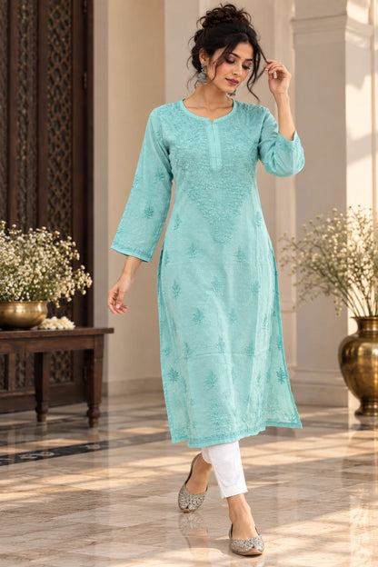 A woman models a light turquoise blue-green embroidered kurta with three-quarter sleeves, paired with white pants and silver embellished flats, standing in a decorated indoor hallway.