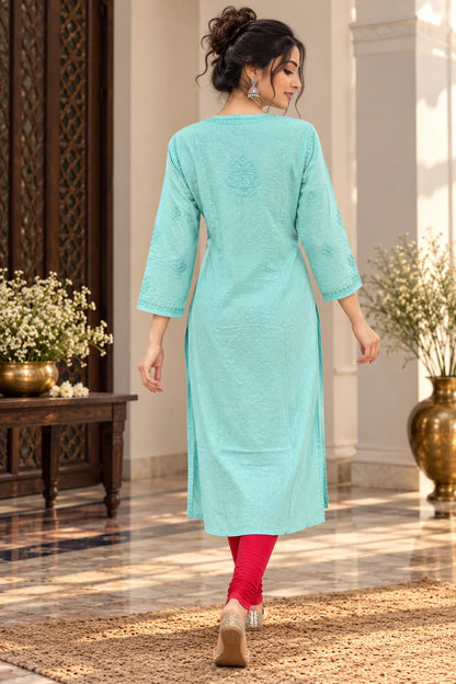 A woman wearing a mint green embroidered kurta with three-quarter sleeves, paired with bright pink leggings and brown heels, walking in a decorated indoor corridor.