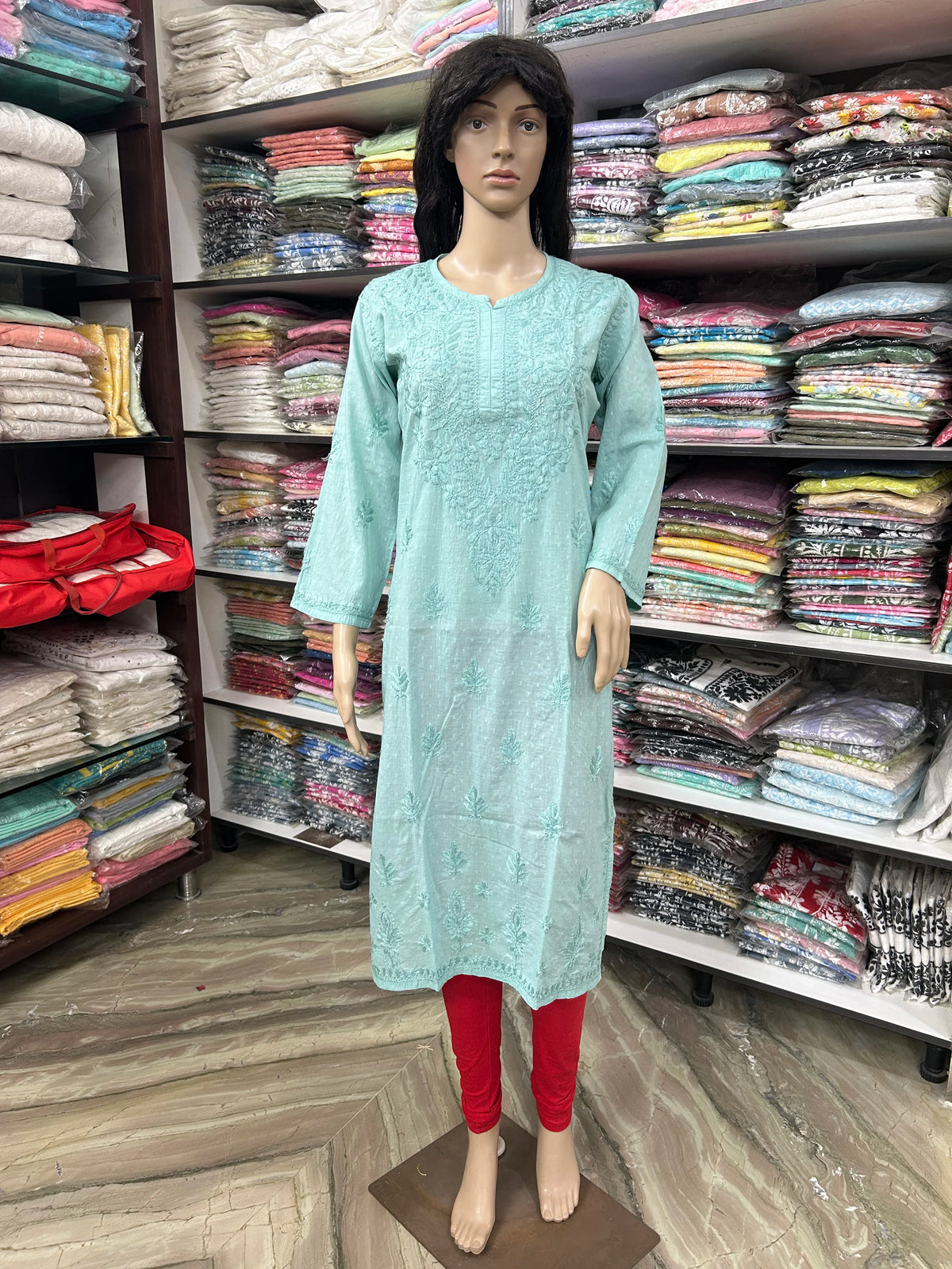 A mannequin wearing a light mint green embroidered kurta with red leggings, standing in a clothing store with shelves of folded garments in the background.