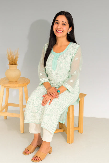 A woman wearing a mint green and white embroidered kurta set sits on a wooden stool next to a small wooden side table with a vase of dried grass, against a light gray studio background.