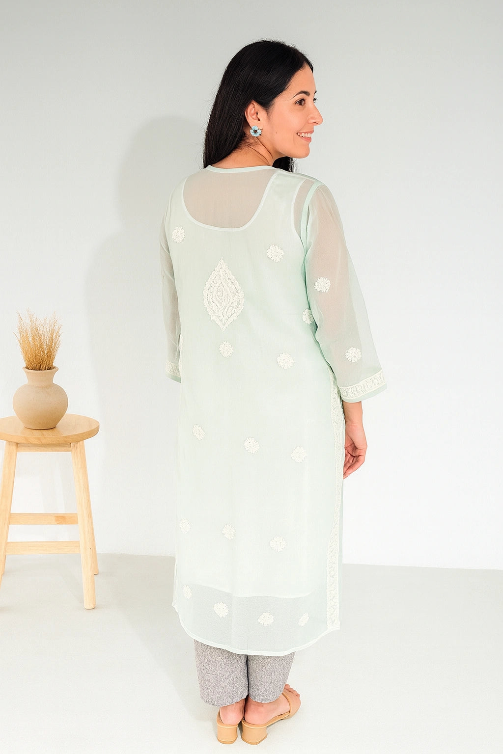 A woman wearing a mint green embroidered kurta with sheer sleeves and white floral embroidery, shown from the back in a studio setting with a small wooden stool on the left.