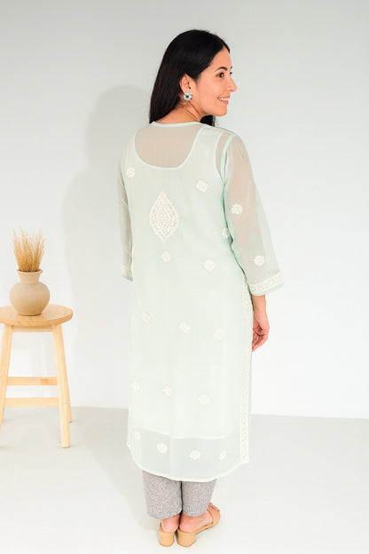 A woman wearing a mint green embroidered kurta with sheer sleeves and white floral embroidery, shown from the back in a studio setting with a small wooden stool on the left.