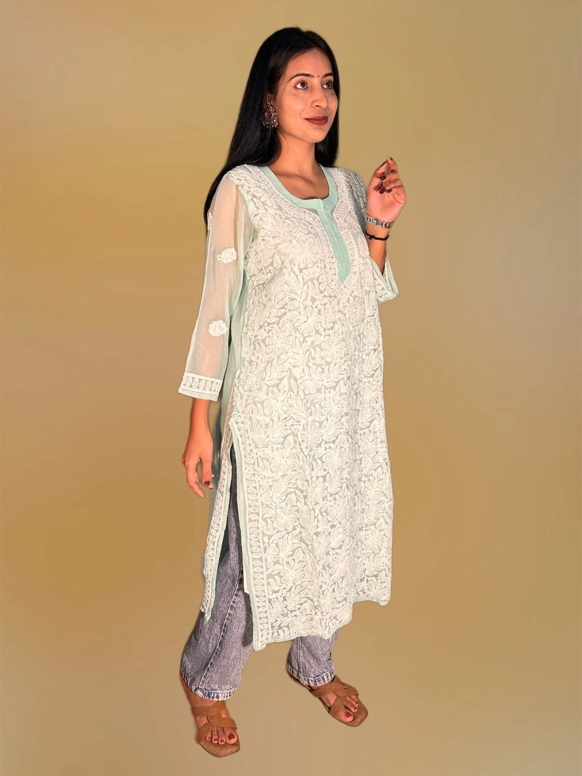 A woman modeling a white embroidered kurta with sheer mint green sleeves, paired with gray patterned pants and brown sandals, against a solid beige background.