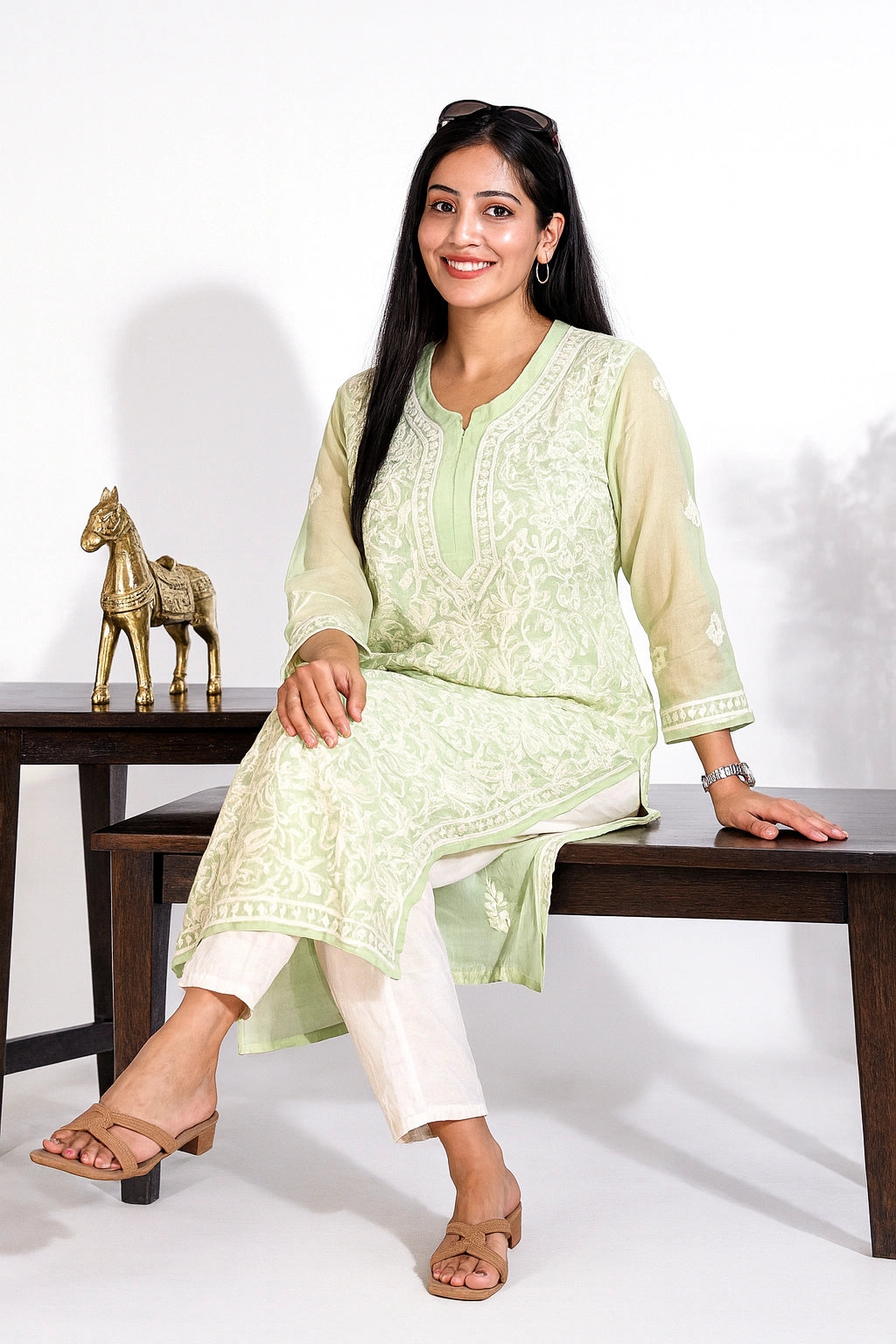 A woman models a light green embroidered kurta with white pants, sitting on a dark wooden bench against a white studio background.