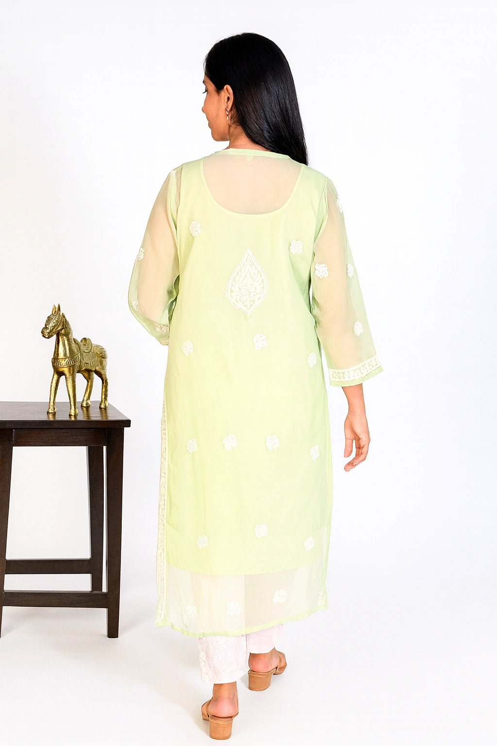 A woman viewed from behind wearing a light yellow-green kurta with white embroidered motifs and sheer sleeves, standing in a studio next to a small table with a gold horse figurine.