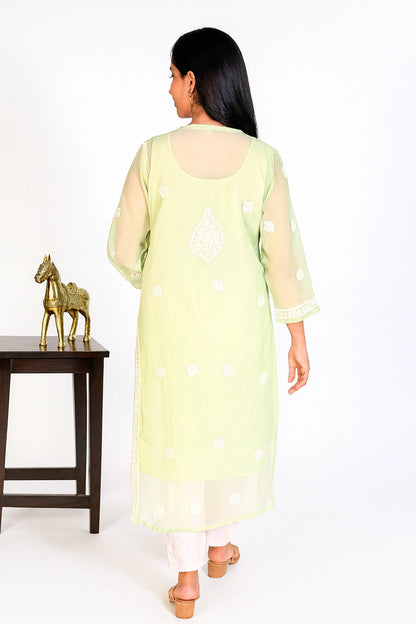 A woman viewed from behind wearing a light yellow-green kurta with white embroidered motifs and sheer sleeves, standing in a studio next to a small table with a gold horse figurine.