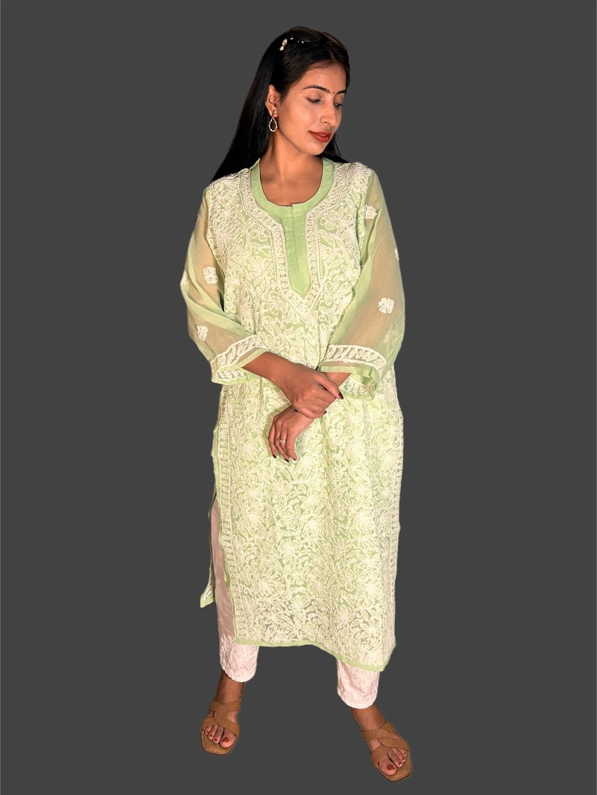 A woman modeling a light green and white embroidered kurta set with sheer sleeves, paired with white pants, against a solid dark gray background.