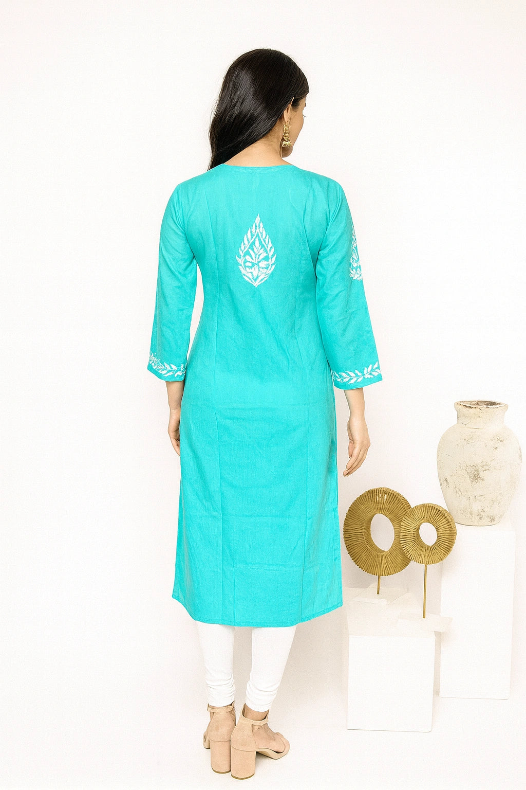 Lucknowi chikankari A woman viewed from the back wearing a turquoise long kurta with white printed motifs on the back and sleeve cuffs, paired with white leggings and beige heels, standing against a white studio background with decorative vases.