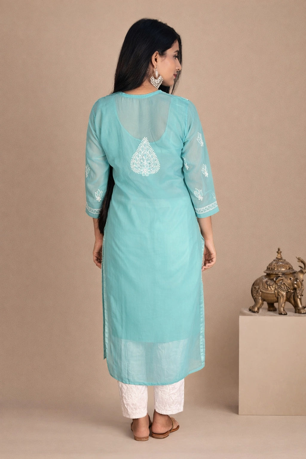 Lucknowi chikankari A woman is shown from the back wearing a light turquoise blue kurta with white embroidery and white pants, standing against a solid beige background.