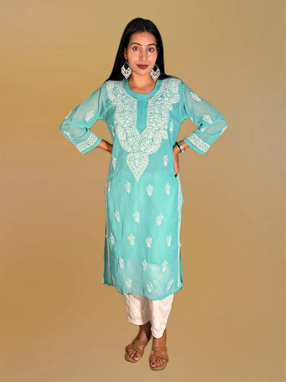 A woman modeling a light turquoise blue kurta with white floral embroidery, paired with white pants and brown sandals, set against a solid beige background.