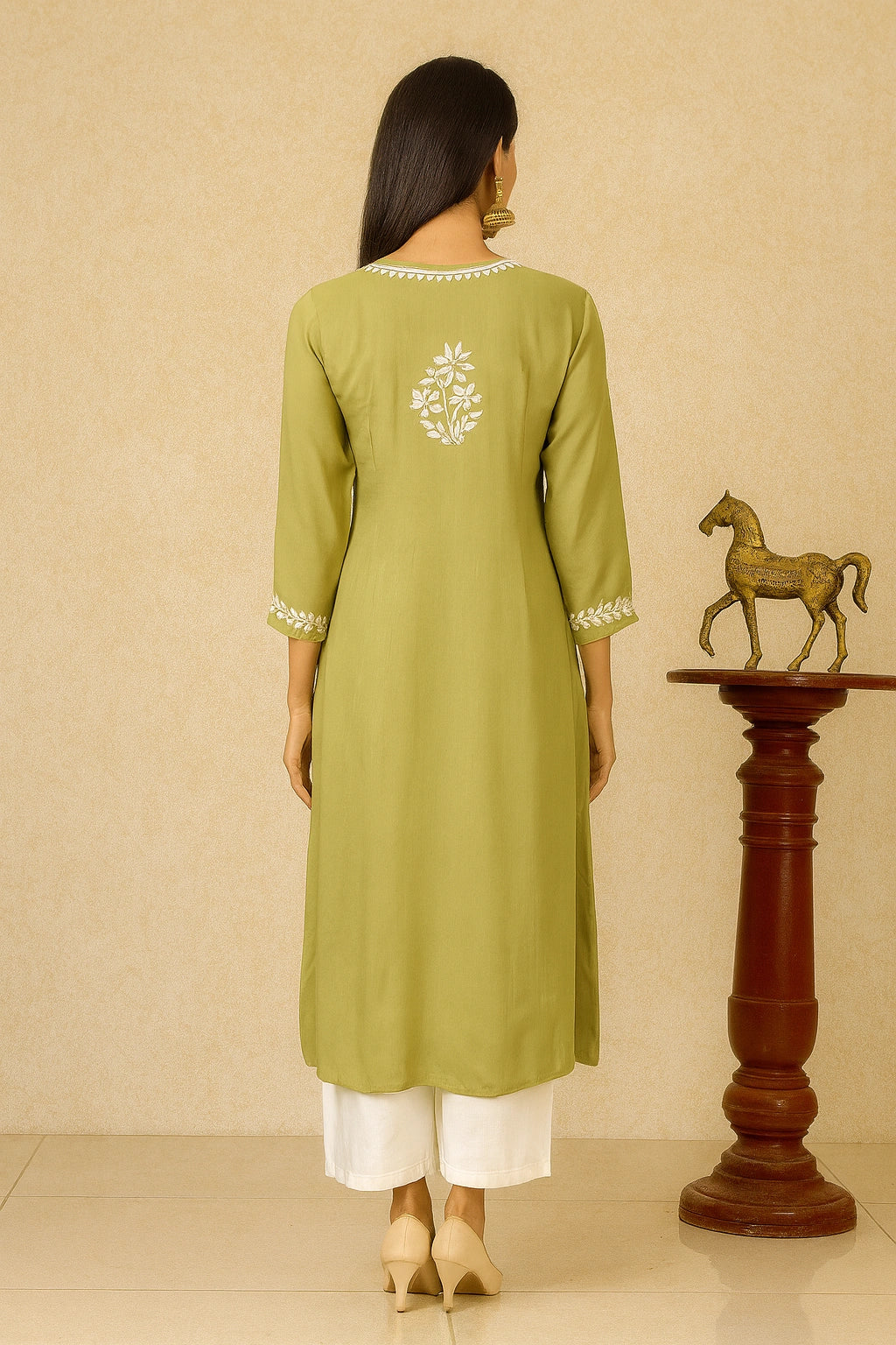 Woman wearing a green kurta with white embroidery, standing against a beige wall with a wooden horse statue.