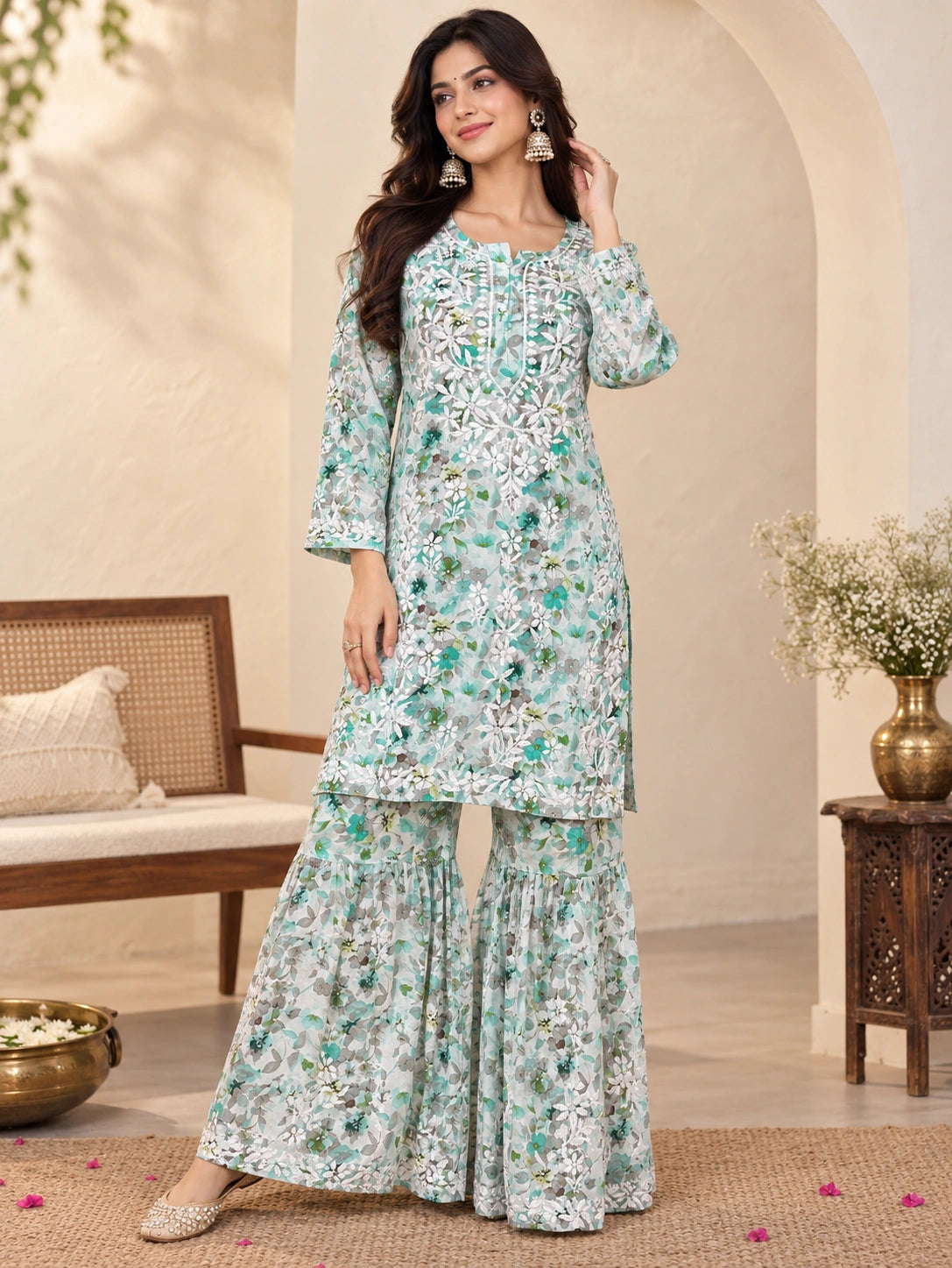 A woman models a light green and white printed salwar kameez suit with intricate embroidery, set against a warm, neutral-toned indoor backdrop with a wooden bench and side table.