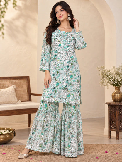 A woman models a light green and white printed salwar kameez suit with intricate embroidery, set against a warm, neutral-toned indoor backdrop with a wooden bench and side table.