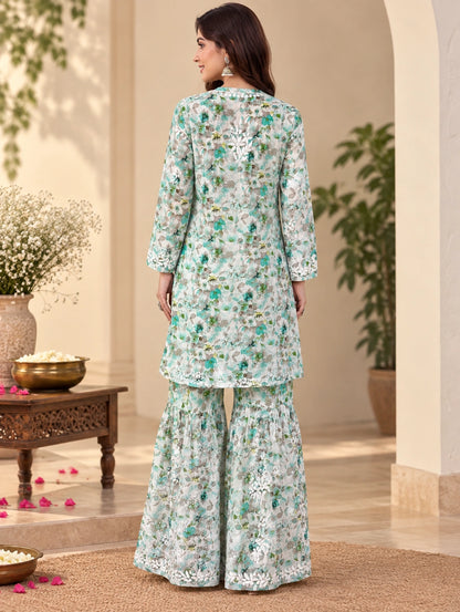 Lucknowi chikankari A woman stands with her back to the camera wearing a light mint green and white floral printed three-piece traditional outfit with wide-legged pants and a long kurta, set against an elegant beige architectural background with potted plants.