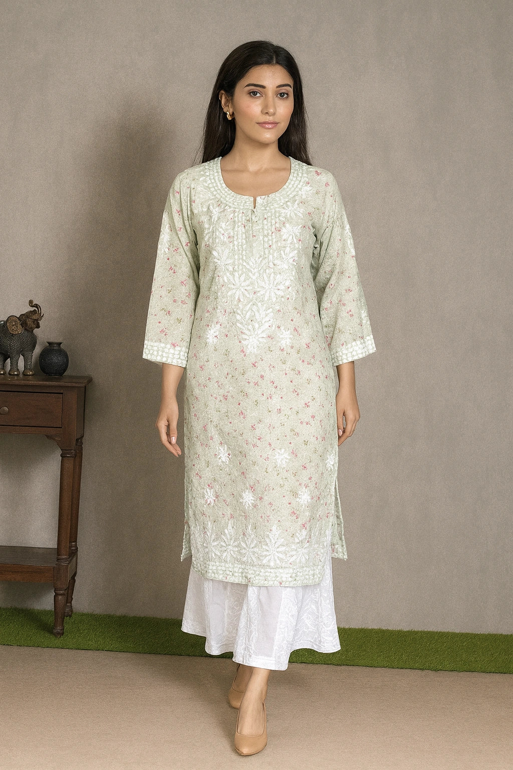 A woman modeling a light sage green kurta with white floral embroidery and a white flared skirt, standing in front of a neutral gray wall.