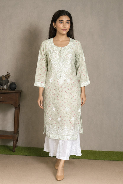 A woman modeling a light sage green kurta with white floral embroidery and a white flared skirt, standing in front of a neutral gray wall.