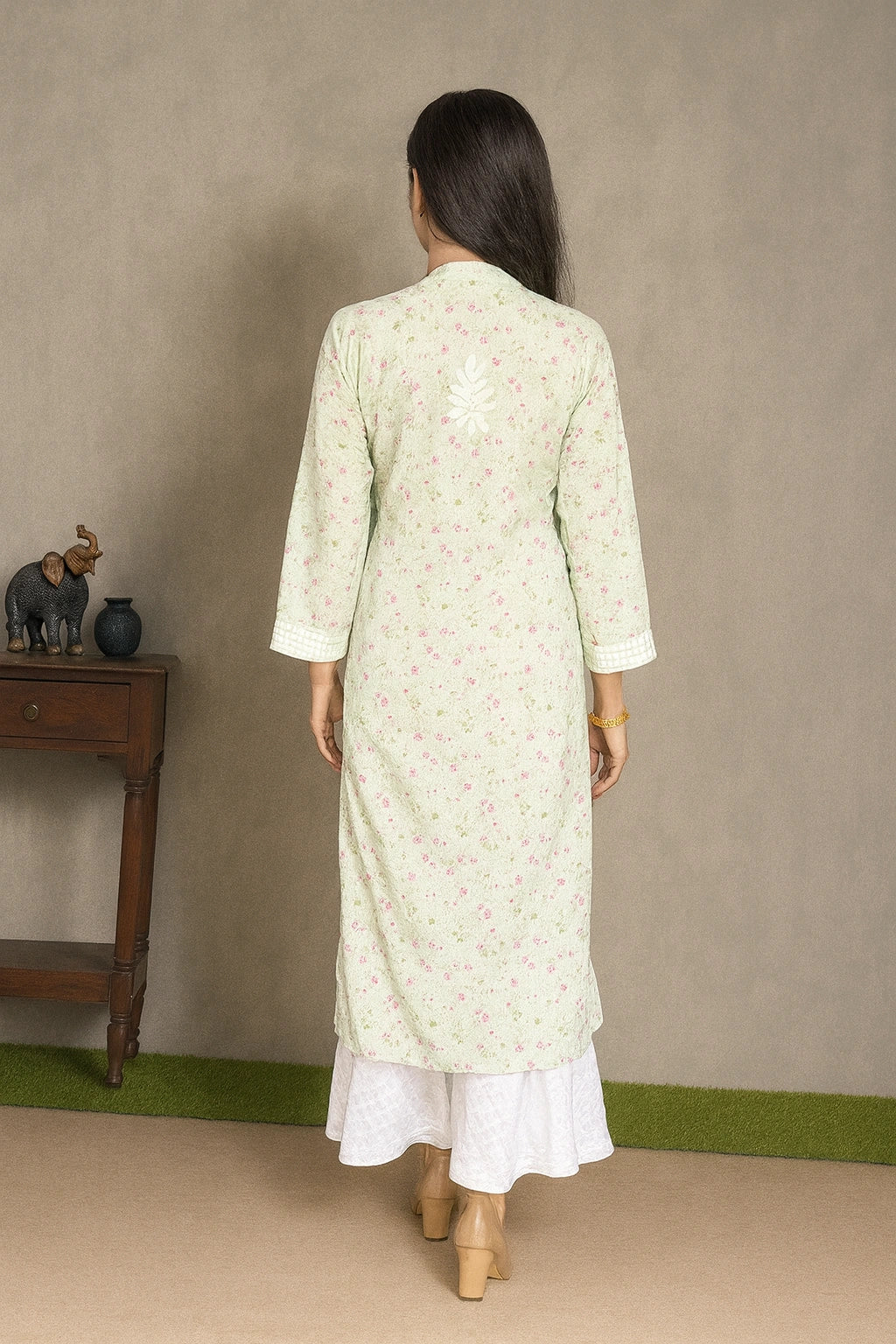 Lucknowi chikankari A woman viewed from behind wearing a light sage green long kurta with a delicate pink floral print, layered over a white skirt. She is standing in front of a neutral gray wall with a small wooden side table on the left.