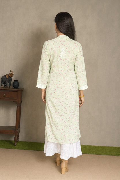 Lucknowi chikankari A woman viewed from behind wearing a light sage green long kurta with a delicate pink floral print, layered over a white skirt. She is standing in front of a neutral gray wall with a small wooden side table on the left.