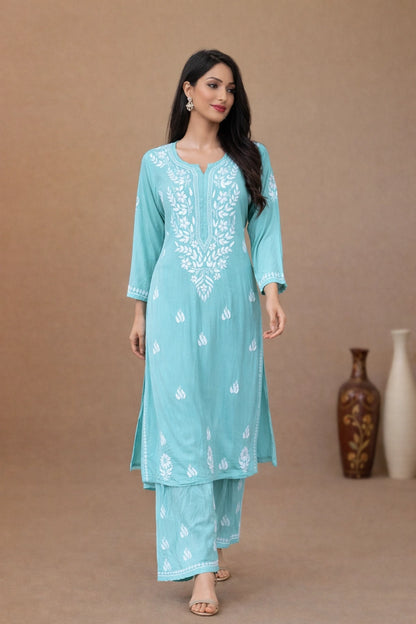 A woman modeling a light blue-green kurta and pant set with white embroidery, standing against a solid brown studio backdrop with decorative vases in the background.