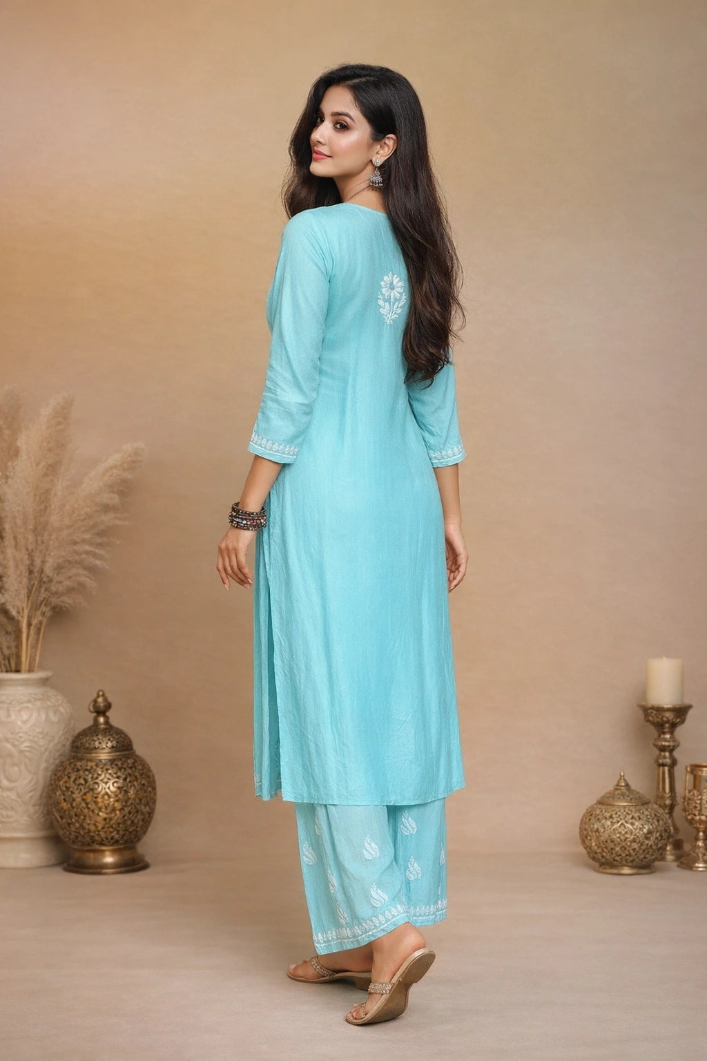 A woman modeling a light blue three-piece salwar kameez suit with white printed motifs and silver embroidery, shown from a back-three-quarter view against a warm beige background with decorative props.
