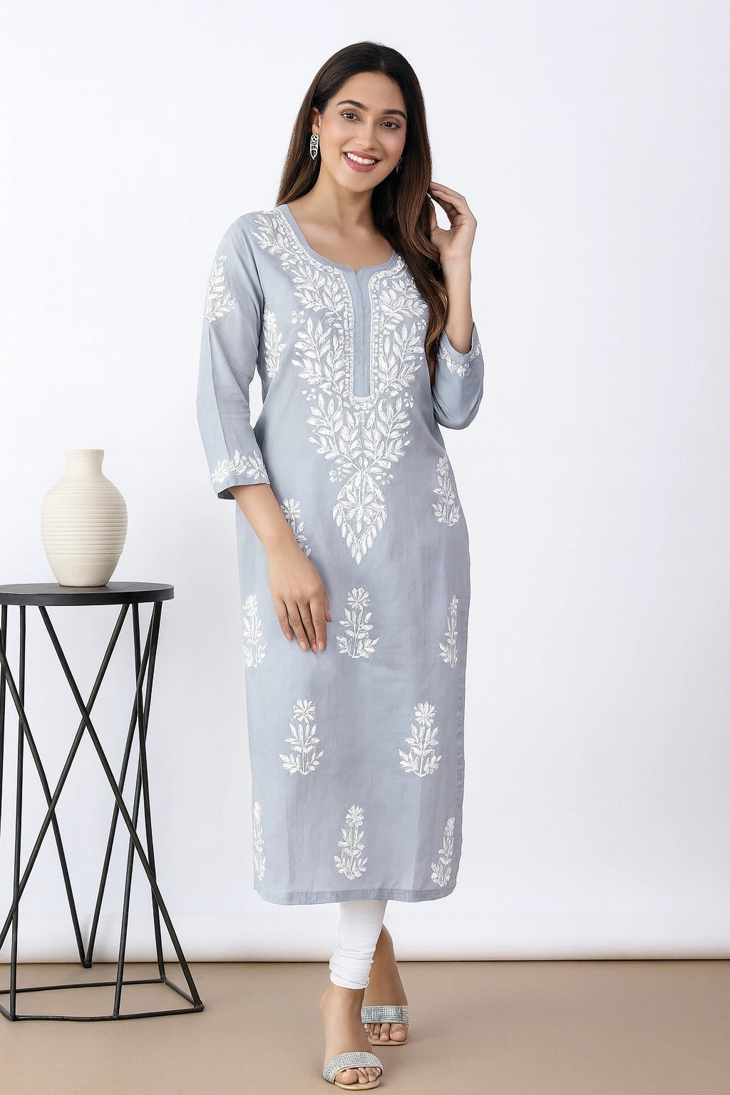 A woman models a light blue-gray three-quarter sleeve kurta with white floral embroidery, paired with white leggings and silver sandals, standing in a studio setting next to a small black side table with a white vase.