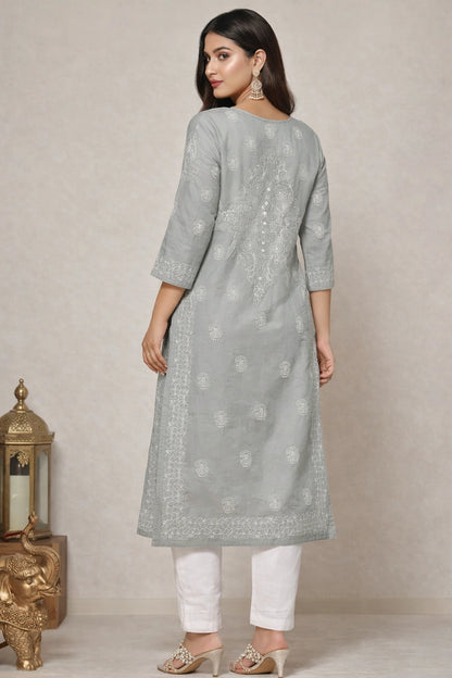 A woman modeling a light gray three-quarter sleeve kurta with white floral embroidery, paired with white pants and beige heels, shown from the back against a neutral beige background.
