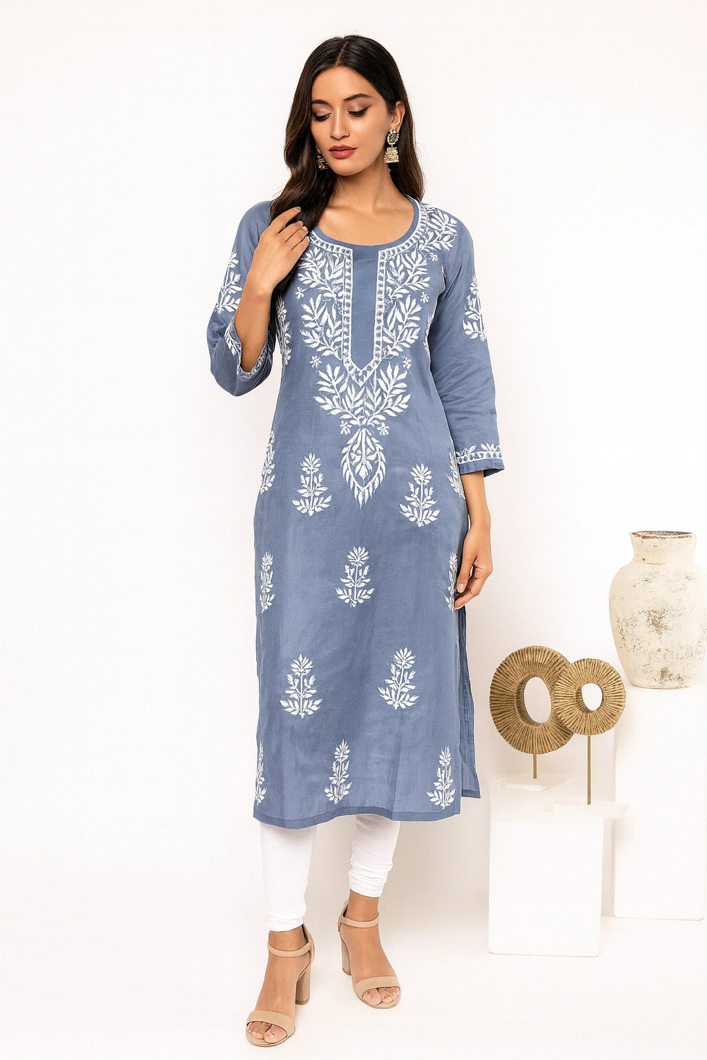 A woman modeling a light sage green kurta with white floral embroidery, paired with white pants and beige sandals, standing in a studio setting.