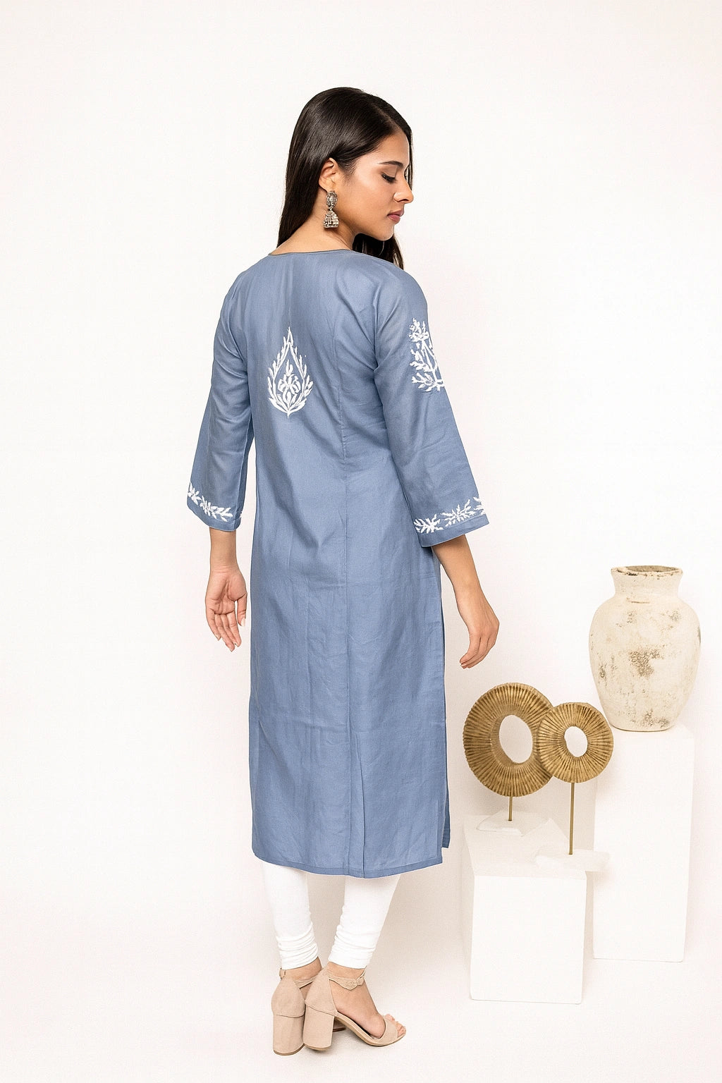 A woman modeling a light gray three-quarter sleeve kurta with white floral embroidery, paired with white pants and beige heels, shown from the back against a neutral beige background.