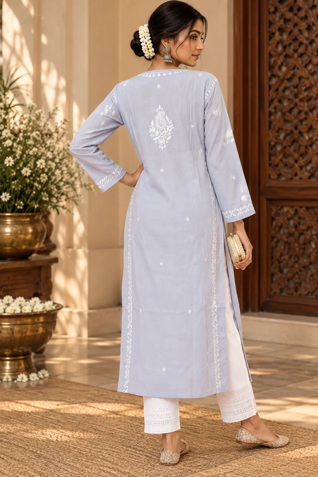 A woman wearing a grey-colored embroidered kurta with white pants, standing in front of a carved wooden door with floral decorations.
